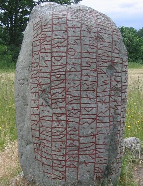 Ein Runenstein zur dekorativen Auflockerung der Seite.
Ein ca. 160cm grauer Stein mit vertikalen Runeninschrifts-Reihen, die geritzt und dann mit roter Farbe gefüllt wurden.